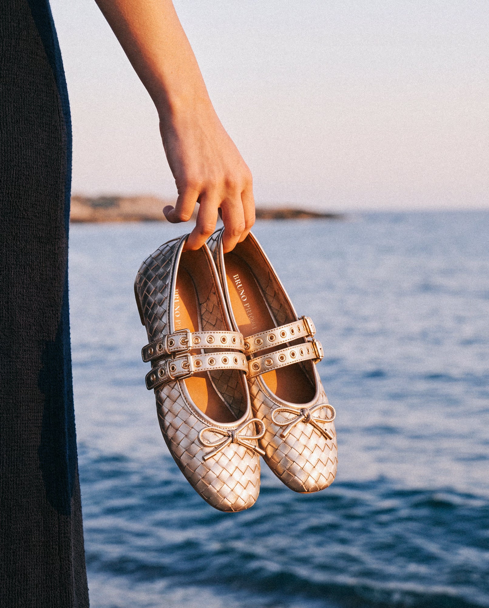 Hand holding a pair of Bruno Premi gold metallic woven leather Mary Jane ballet flats with double buckle straps, eyelet detail and bow toe, against a blurred sea and coastline background.