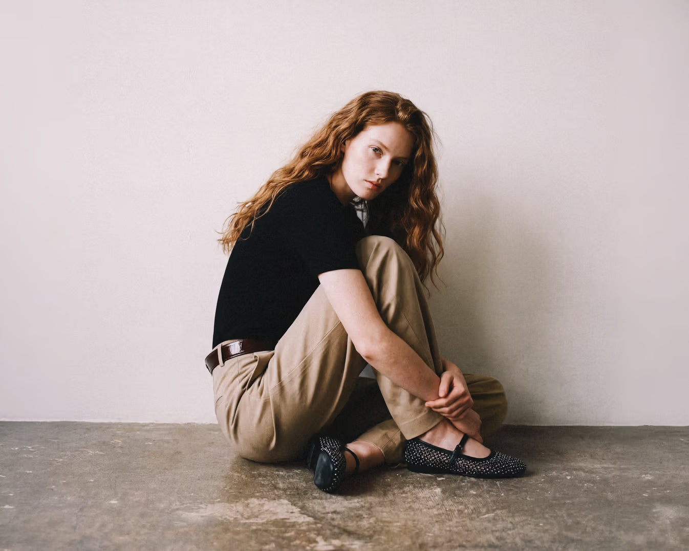 Woman sitting on a concrete floor wearing black mesh Mary Jane ballet flats by Bruno Premi, styled with khaki trousers, a black t-shirt and a brown leather belt. Minimal light grey background.