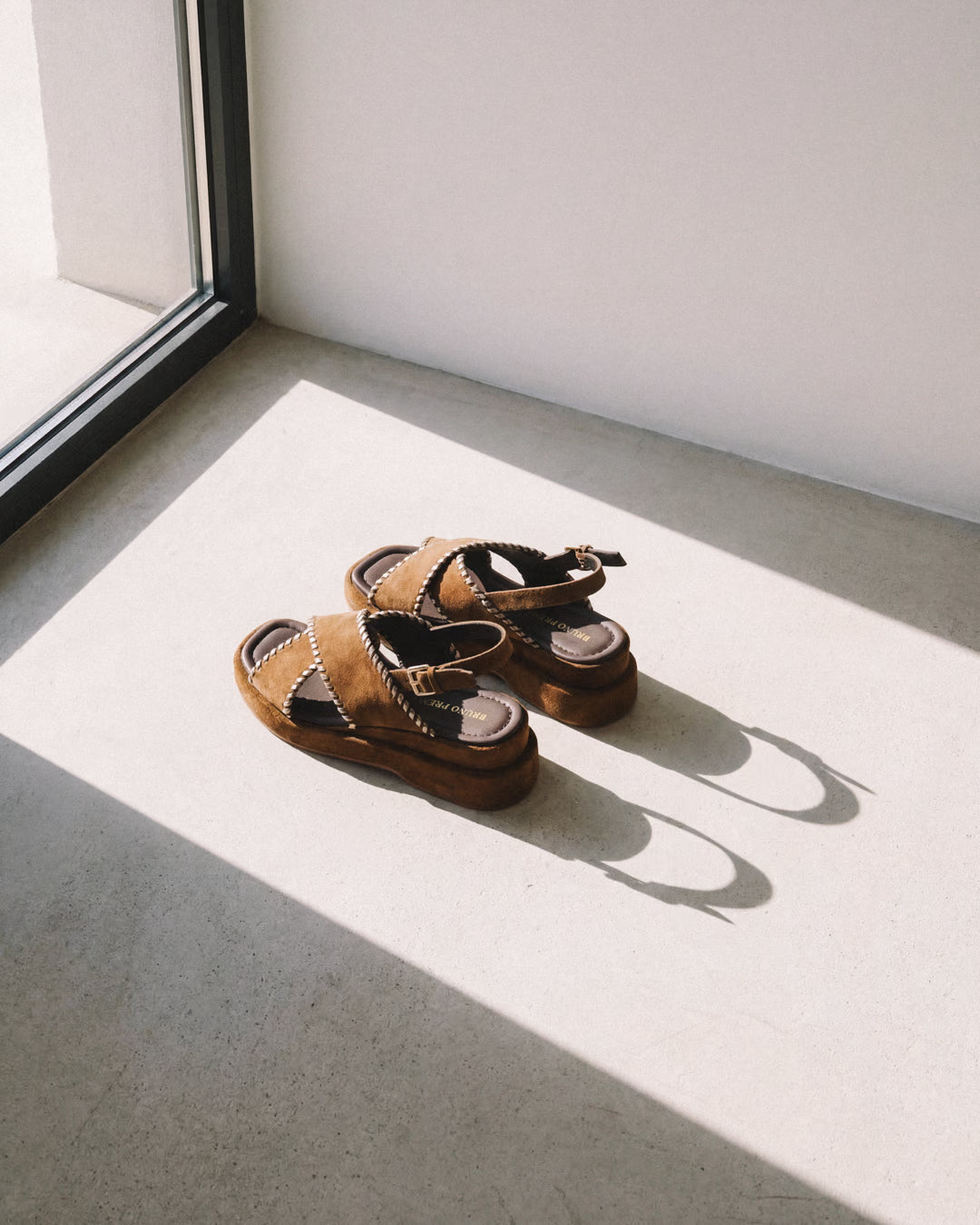 Bruno Premi brown suede platform sandals with black crossover straps and contrast stitching detail, placed on a concrete floor in natural window light.